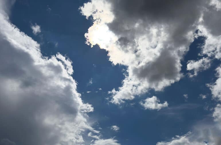 Partly cloudy sky with large, fluffy clouds partially covering the sun, creating bright light and shadows against a deep blue background.